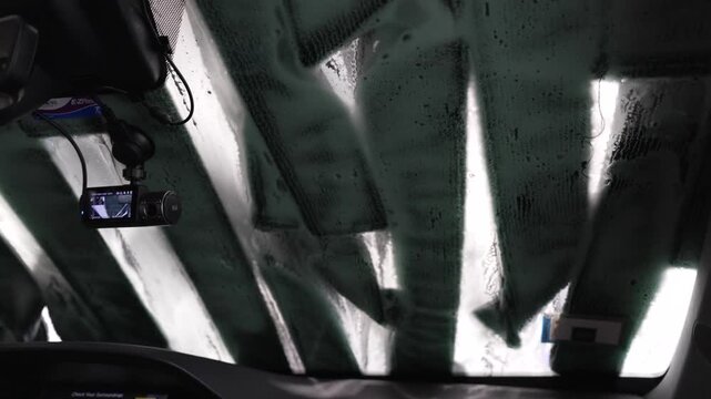 A professional 4K POV video shot from inside a car driving through an automatic tunnel car wash. Thick white soap foam, water jets, and spinning brushes create dynamic patterns and textures on the win