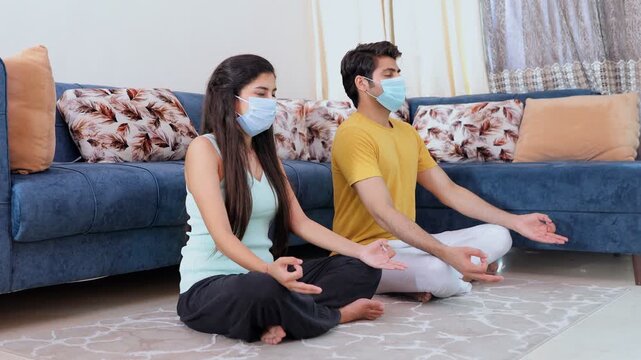 Young boyfriend girlfriend meditating in Sukhasana (easy pose) for a healthy lifestyle. Sick Indian couple doing breathing exercises while wearing surgical masks during the coronavirus/Covid-19 pan...
