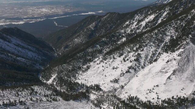 Drone footage of snow covered mountain slopes and conifer forest above a deep valley near Galina Peak, Arshan, Republic of Buryatia, Russia on a clear winter day
