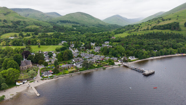 Aerial view of the historic Luss village on the shore of Loch Lomond, showcasing traditional stone cottages, a pier, a church, and lush green mountains in the Scottish Highlands.