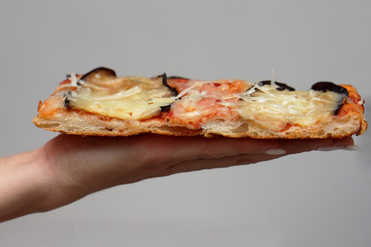 Side profile view of a thick, porous rectangular slice of pizza parmigiana held on a female hand against a plain grey background