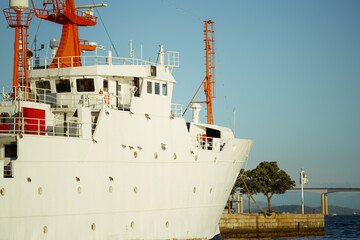 Naklejka premium Telephoto view of a white ship bow and bridge with orange masts moored beside a pier, water and a long bridge visible in warm sunlight