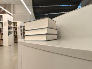 Fototapeta premium Stack of three closed books resting on a curved white table in a modern library with shelves of books and a staircase visible in the background