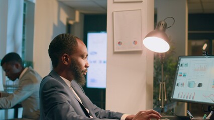 Fototapeta premium African american businessmen sitting at desk, looking over financial charts, imputing data on computer during nightshift. Coworkers solving company project tasks overnight before deadline