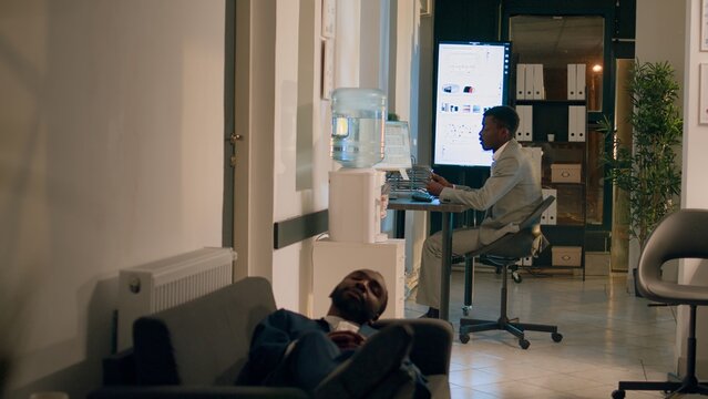 Manager finishing nightshift, ready to go home, catching employee taking nap on office couch. Management executive annoyed by worker slacking off, sleeping at work, thinking of firing him