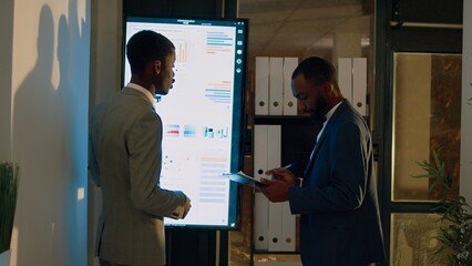 Fototapeta premium Manager debriefing employee in front of digital screen, showing him important research statistics before getting back to work. Businessmen in office during nightshift brainstorming business insights