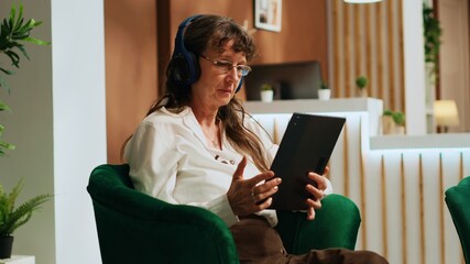 Retired woman using tablet in lobby, watches movie on streaming services while she waits to see room reservation. Old person enjoying modern technology with device and headset. Handheld shot. © DC Studio