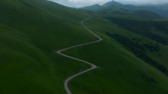 Aerial footage of a winding road crossing lush green mountain slopes in Aktoprak, Kabardino-Balkaria, Russia, with misty peaks and dramatic cloudy weather