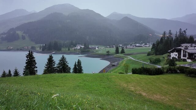 Rainy day over Davos alpine lake and mountain village with green meadows, pine trees and chalets in misty foggy weather, Switzerland cinematic video