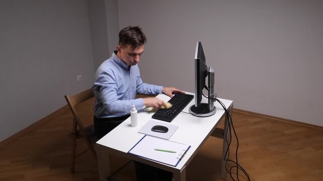 Man in a blue shirt carefully sanitizing a computer keyboard with a cloth and spray bottle on a white desk, maintaining a clean, hygienic office or home workspace