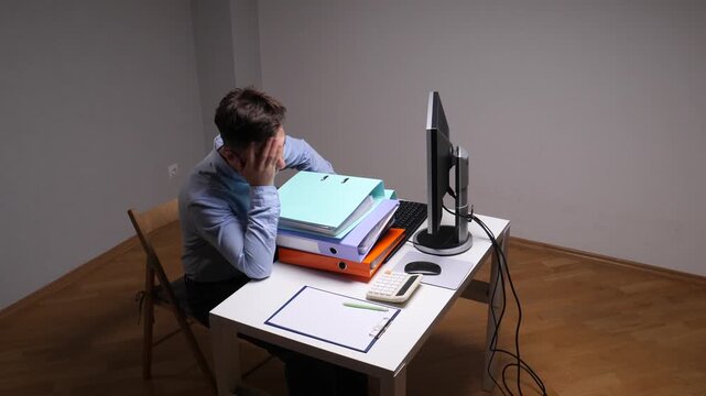 Tired office worker slumped at desk surrounded by binders and papers, struggling with overwhelming workload, fatigue and stress while working late on computer