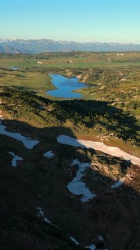 Vertical Screen: Beartooth Highway in Montana USA reveals stunning alpine lake nestled among evergreen forests with melting snow patches on rocky terrain and distant mountain peaks under clear blue.