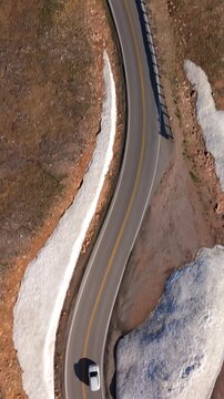 Vertical Screen: Beartooth Highway in Montana USA with white car navigating curved mountain road. Drone footage captures winding asphalt through rocky alpine terrain with snow patches and guardrails.
