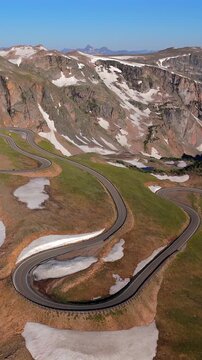 Vertical Screen: Beartooth Highway in Montana, USA curves through alpine terrain with snow patches on green meadows. Rugged peaks with melting snow rise dramatically under clear blue sky.