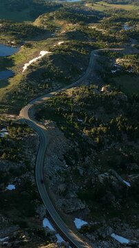 Vertical Screen: Beartooth Highway curves through Montana USA alpine landscape with scattered lakes and evergreen forests. Golden hour sunlight illuminates rocky terrain and snow patches.