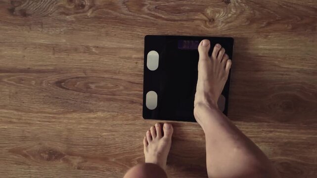 POV - top point of view video from above: woman bare feet stepping on black digital floor scales, she weighing, checking herself at home. Measuring weight, control, wellness, diet body care concept