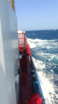 Vertical shot of a cargo ship sailing through rough sea waves on a sunny day.
