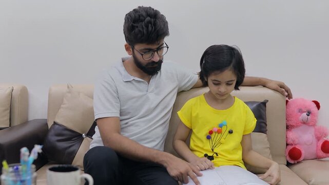 Indian father checking his school-age daughter's homework in a notebook. Medium shot of a young dad helping his little child in studies while sitting on a sofa - education 