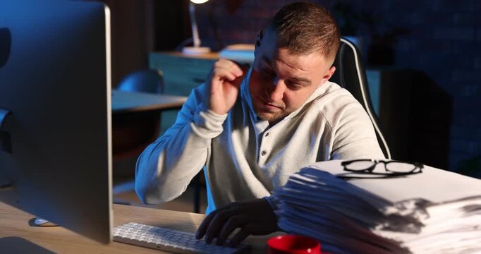 Workaholic. Tired man working with computer near stack of documents and cup at desk indoors at night. Camera moving forward
