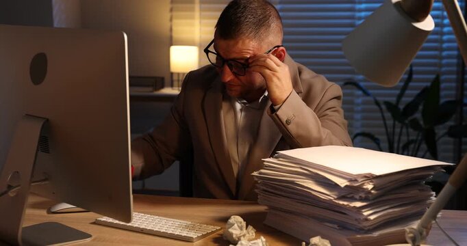 Workaholic. Man checking empty coffee cup at wooden desk with computer and stack of documents indoors at night. Camera moving forward