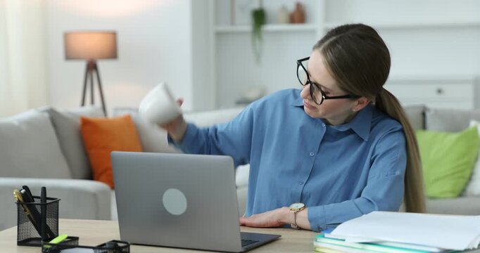 Workaholic. Woman in glasses checking empty coffee cup at wooden desk with computer and stack of documents indoors