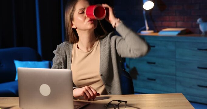 Workaholic. Woman checking empty coffee cup at wooden desk with laptop indoors at night