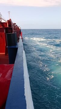 Vertical shot from a cargo ship deck sailing through deep blue sea water on a partly cloudy day.