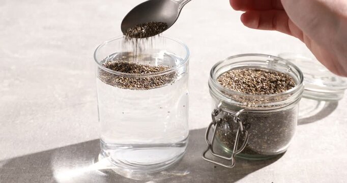 Woman pouring chia seeds into glass of water at light grey table, closeup
