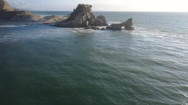 Aerial shot of a lone surfer riding the deep blue waters of La Ti&ntilde;osa beach, with a rocky background and the Santa Marianita lighthouse and beach on the horizon