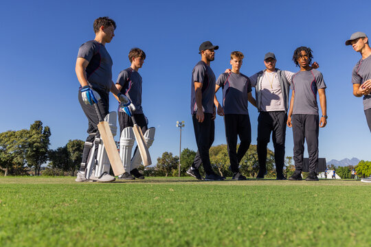 Diverse male cricket team standing on turf pitch holding bats and wearing team shirts