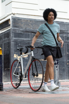 African-American man walking white bike red rim on sidewalk wearing black crossbody bag, copy space