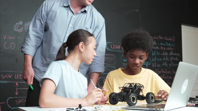 Caucasian teacher pointing at laptop screen while explain car model system at blackboard with coding or programming code written. Happy diverse student learning about robotic construction. Edification