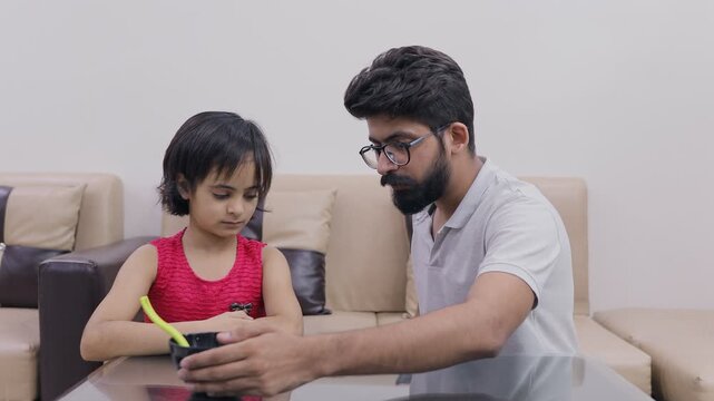 An Indian father in the living room is upset with his daughter for not eating food . Medium shot of a stubborn Indian child refusing to eat healthy food - Angry parent scolding her daughter 