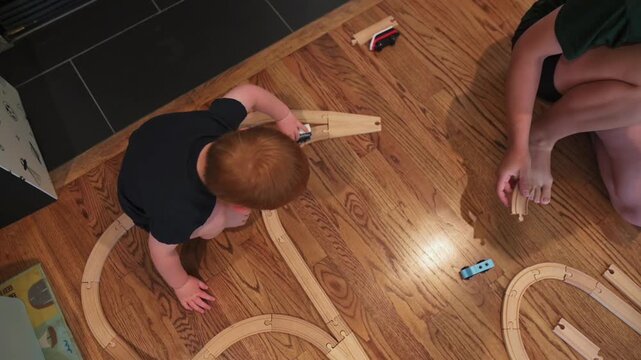 Young boy with red hair interacts with wooden train tracks, carefully placing pieces and maneuvering toy trains along the layout on a wooden floor in a warm indoor play space