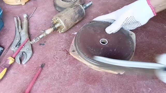A mechanic wearing white gloves vigorously cleans the rust and dirt off the front endshield of a water pump using a stiff wire brush.