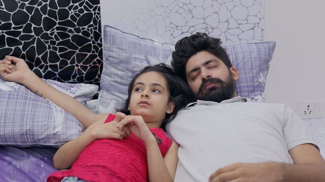 A young dad falls fast asleep on the bed while his little girl talks to him. Medium shot of a tired father sleeping while his young daughter is chatting with him - Caring daughter 