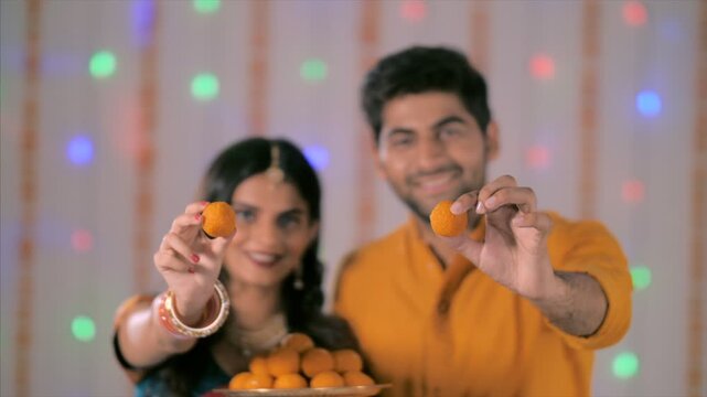 A beautiful newly married Indian couple celebrating Diwali and offering besan ladoo. Happy Indian couple in traditional dressed holding a plate of boondi laddoo and offering them - Diwali Concept