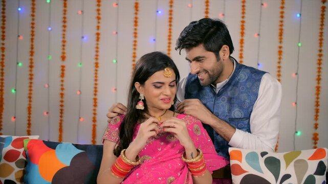 Newly Married Indian husband surprises wife with a necklace as a gift - Festival concept. Handsome husband gives a wonderful necklace as a surprise to his beautiful wife in pink saree - Diwali 