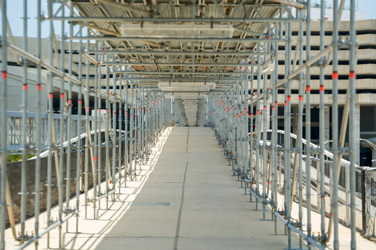 Empty outdoor pedestrian concrete walkway covered with heavy metal construction scaffolding leading towards a parking garage.