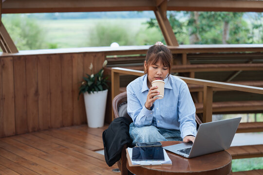 Woman is engaged in financial planning and data analysis while having discussion over coffee using laptop and tablet in wooden outdoor setting with natural light and greenery