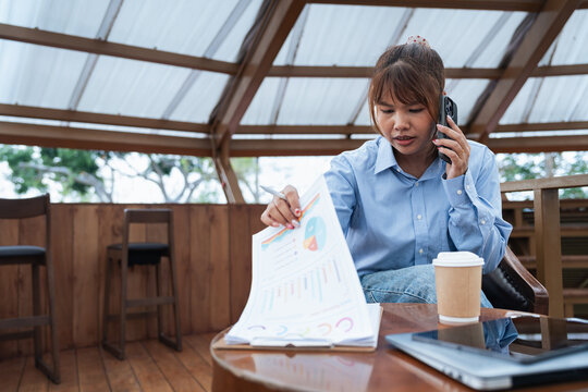 Woman is engaged in financial planning and discussion while analyzing data charts on paper during phone call in wooden indoor setting with laptop and coffee cup nearby