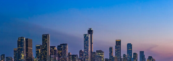 Naklejka premium A wide panoramic evening view of illuminated skyscrapers in the Toronto financial center rising above a dark treeline and highway during a purple twilight dusk in Ontario Canada