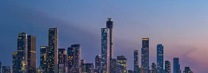Naklejka premium A wide panoramic evening view of illuminated skyscrapers in the Toronto financial center rising above a dark treeline and highway during a purple twilight dusk in Ontario Canada