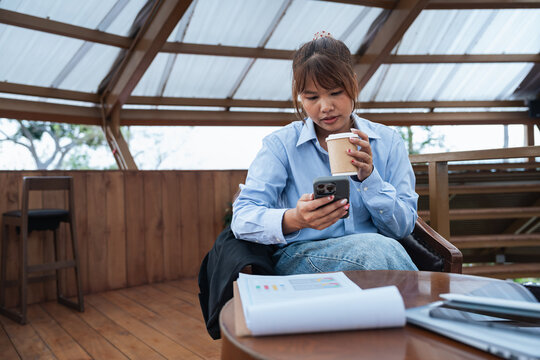 Young woman holding coffee cup and smartphone sitting in wooden room with financial data and analysis documents on table planning discussion