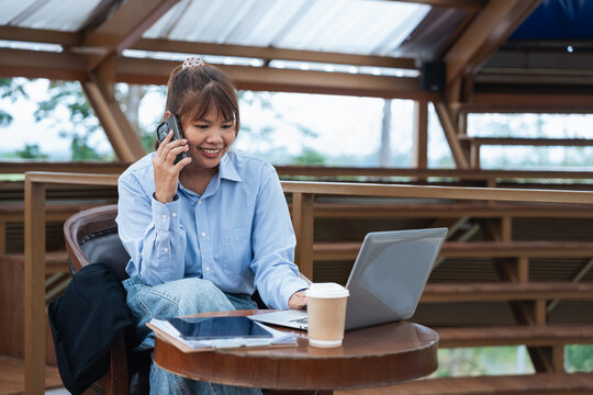 Young woman using laptop and smartphone for financial planning and data analysis while having discussion in cozy outdoor workspace with coffee and tablet on table