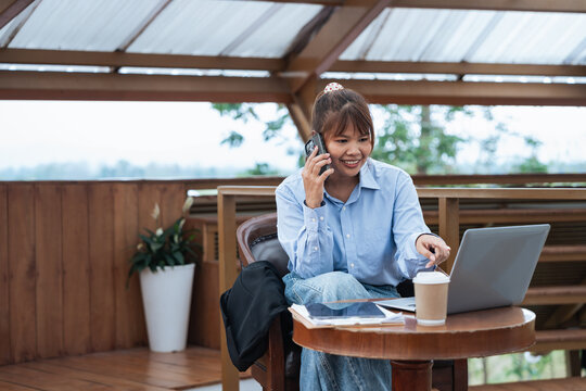 Young woman using laptop and smartphone for financial planning and data analysis while having discussion in cozy wooden terrace with natural light and greenery outside