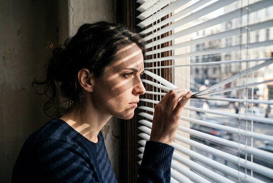 Woman cautiously peeking through blinds looking outside window