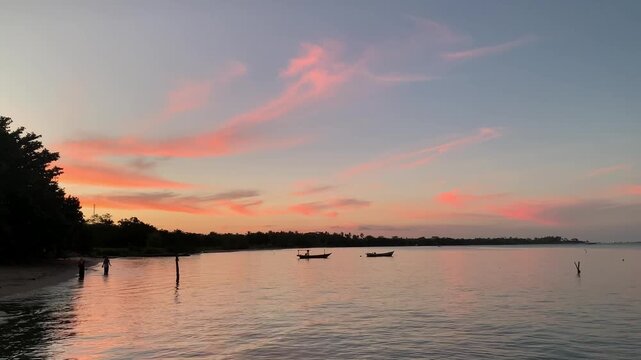 4K sunset seascape at Tanjung Lesung Beach, Indonesia, with two tourist silhouettes photographing by the shore and fishing boat silhouettes. Wide view, calm waves, peaceful travel theme.