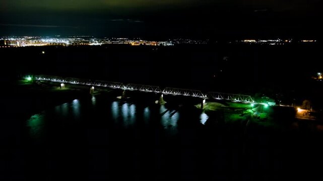 Ufa, Russia. Railway bridge across the Belaya River at night, steel truss structure illuminated with white and green lights reflecting on dark water, panoramic Drone footage