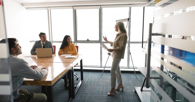 Confident older mature business leader woman holding training seminar, presenting project, marketing strategy, standing at meeting table in boardroom, talking to diverse team. Doorway full length view